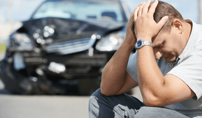 A man sits beside a damaged car on the ground.